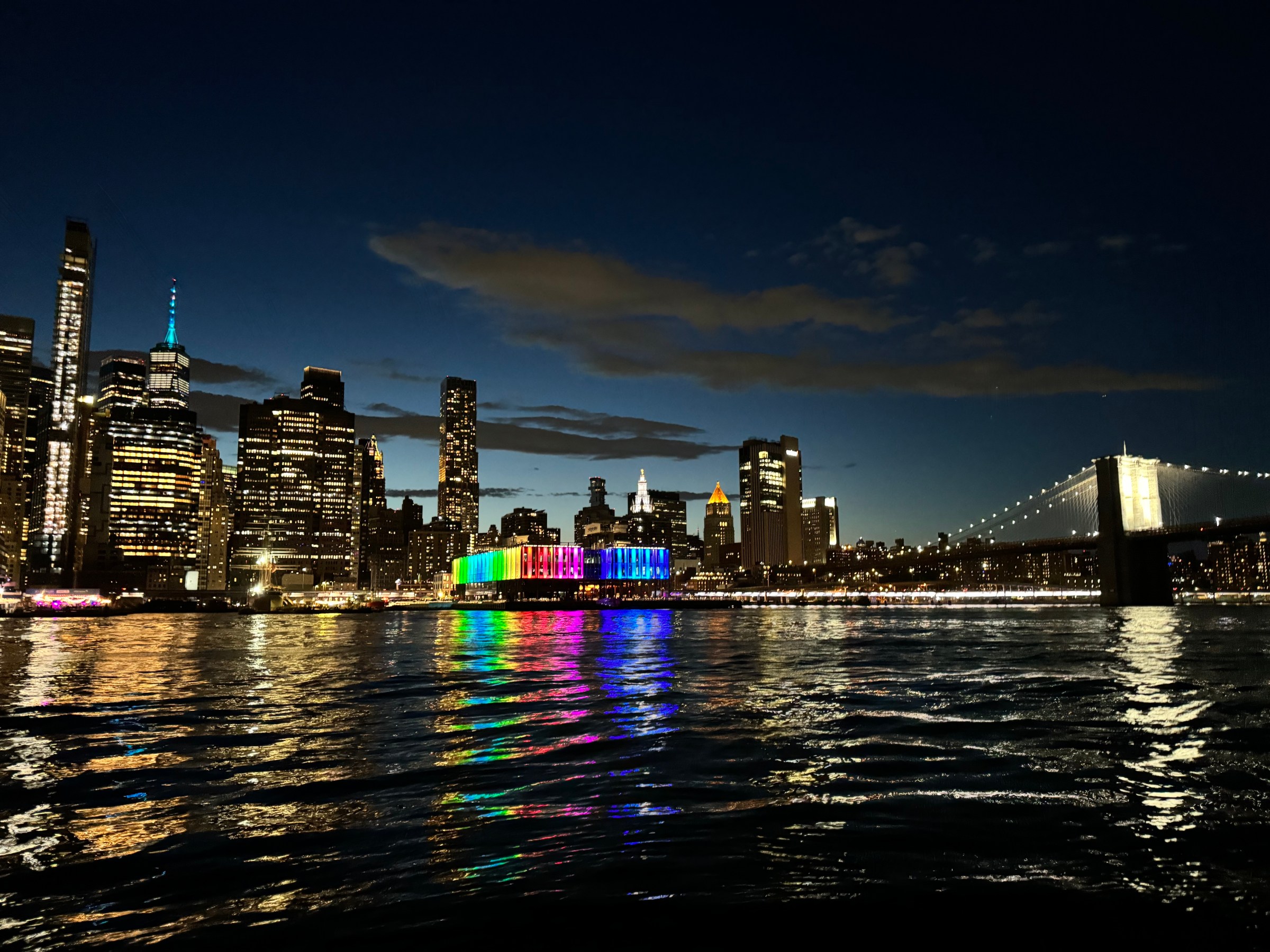City skyline at night with colorful lights reflected in water and a lit bridge on the right.