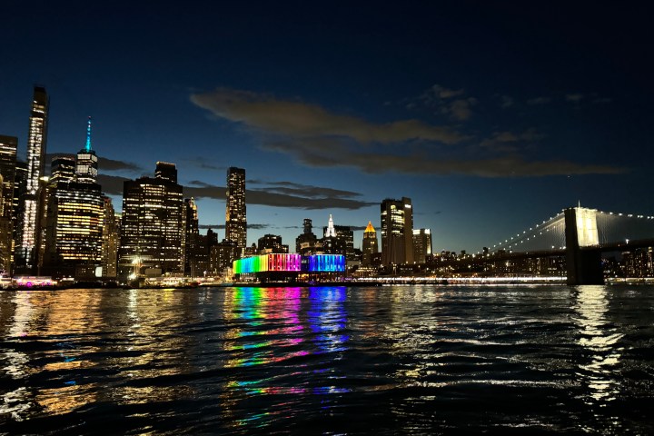 City skyline at night with colorful lights reflected in water and a lit bridge on the right.