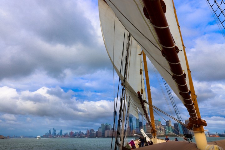 Sailing ship with large sails in foreground, city skyline and cloudy sky in background.