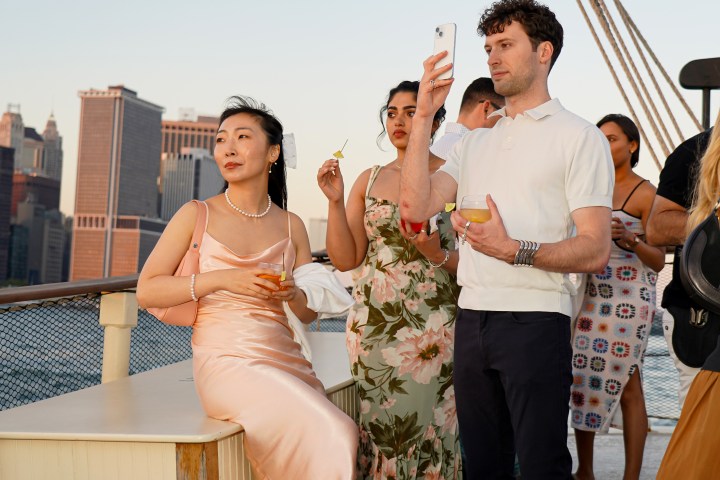 People on a boat deck, dressed elegantly, with a city skyline in the background during sunset.