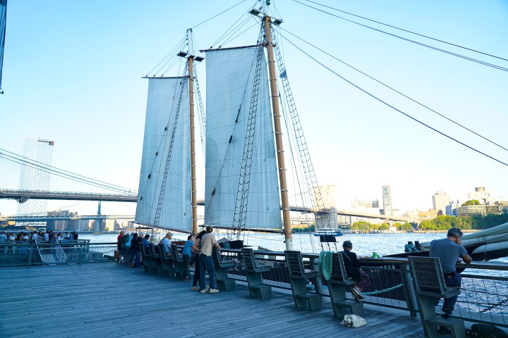 People sitting on benches near a sailboat and a bridge over a city river.