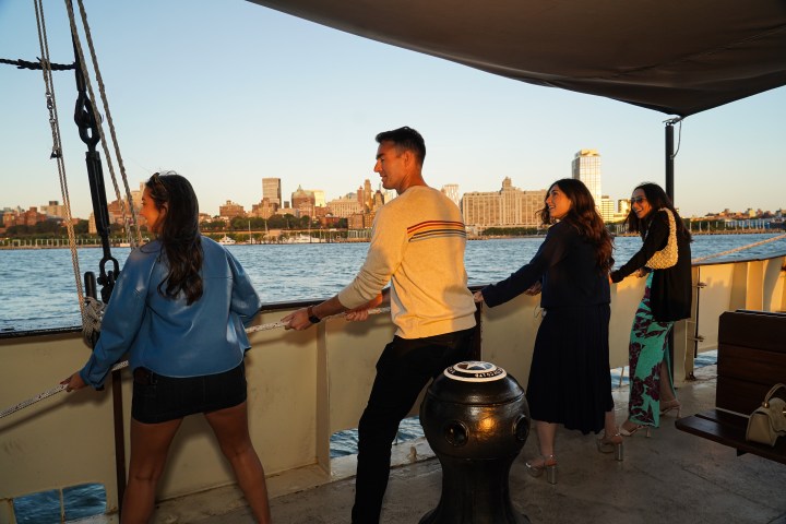 Group of four people on a boat, pulling ropes with city skyline in the background.