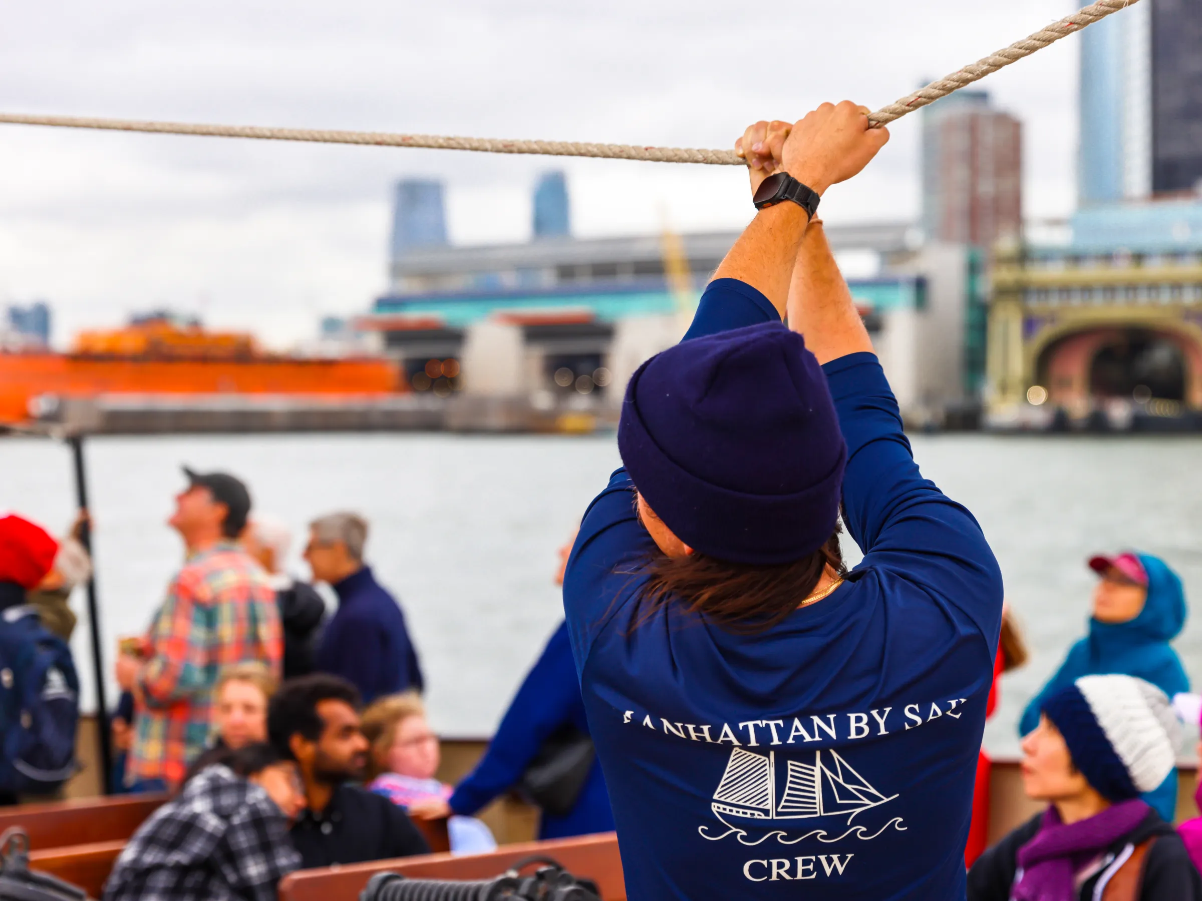 Person in crew shirt pulling rope on a boat, with people watching and cityscape in the background.