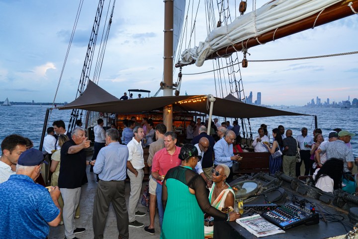 People socializing on a sailboat deck with city skyline at dusk.