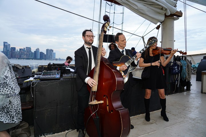 Musicians playing on a boat deck with cityscape in background.
