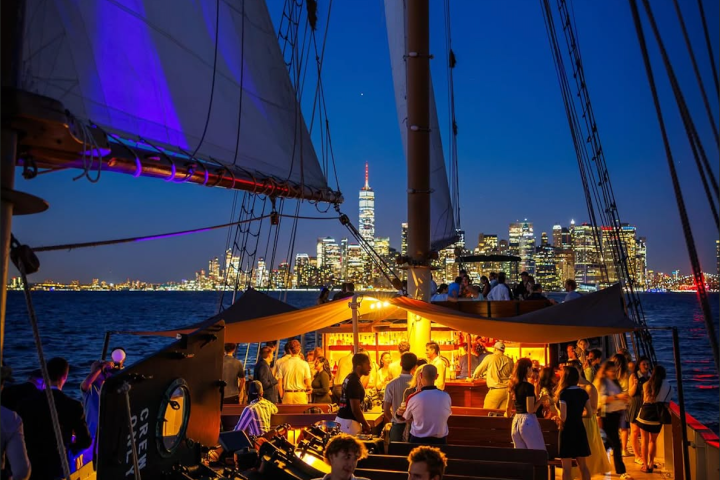 People socializing on a sailboat with city skyline in the background at dusk.