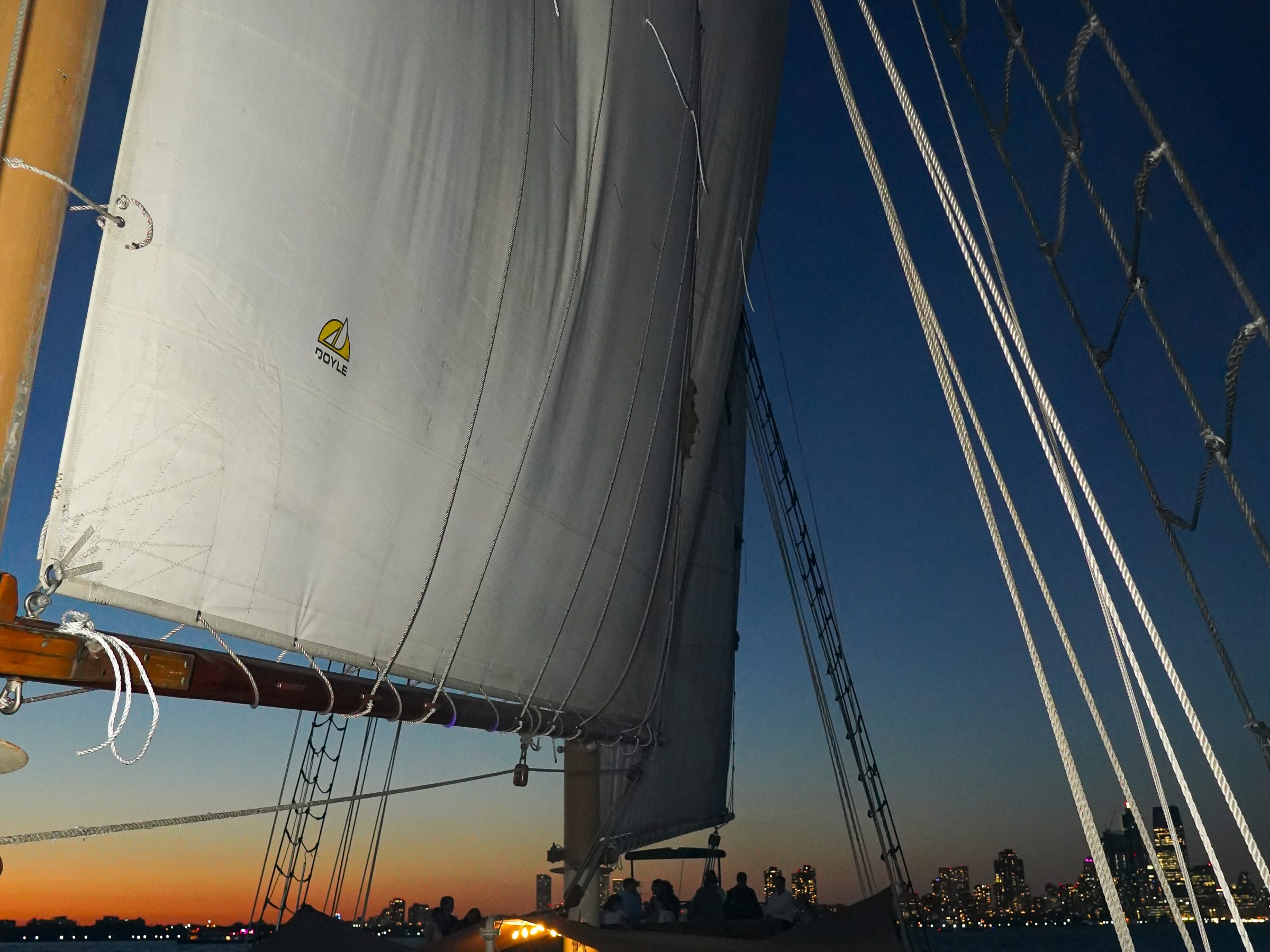 Large sailboat with partygoers on deck at sunset, city skyline visible in the background.