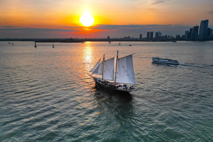 Sunset over water with a sailing ship and distant city skyline.