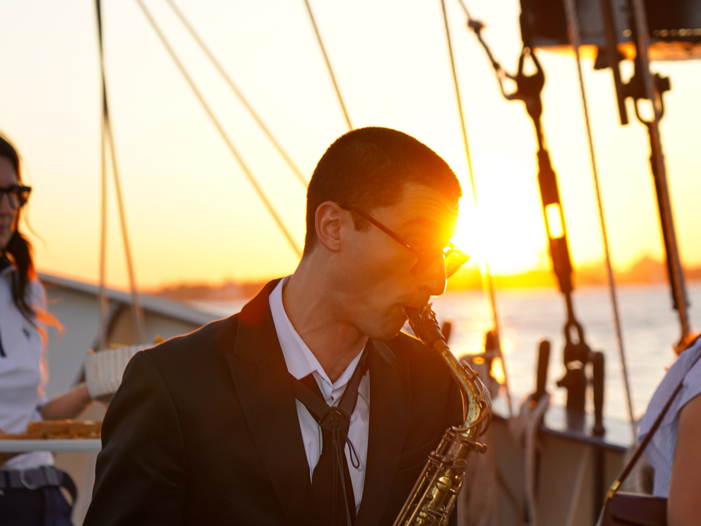 Man in a suit plays saxophone on a boat at sunset.