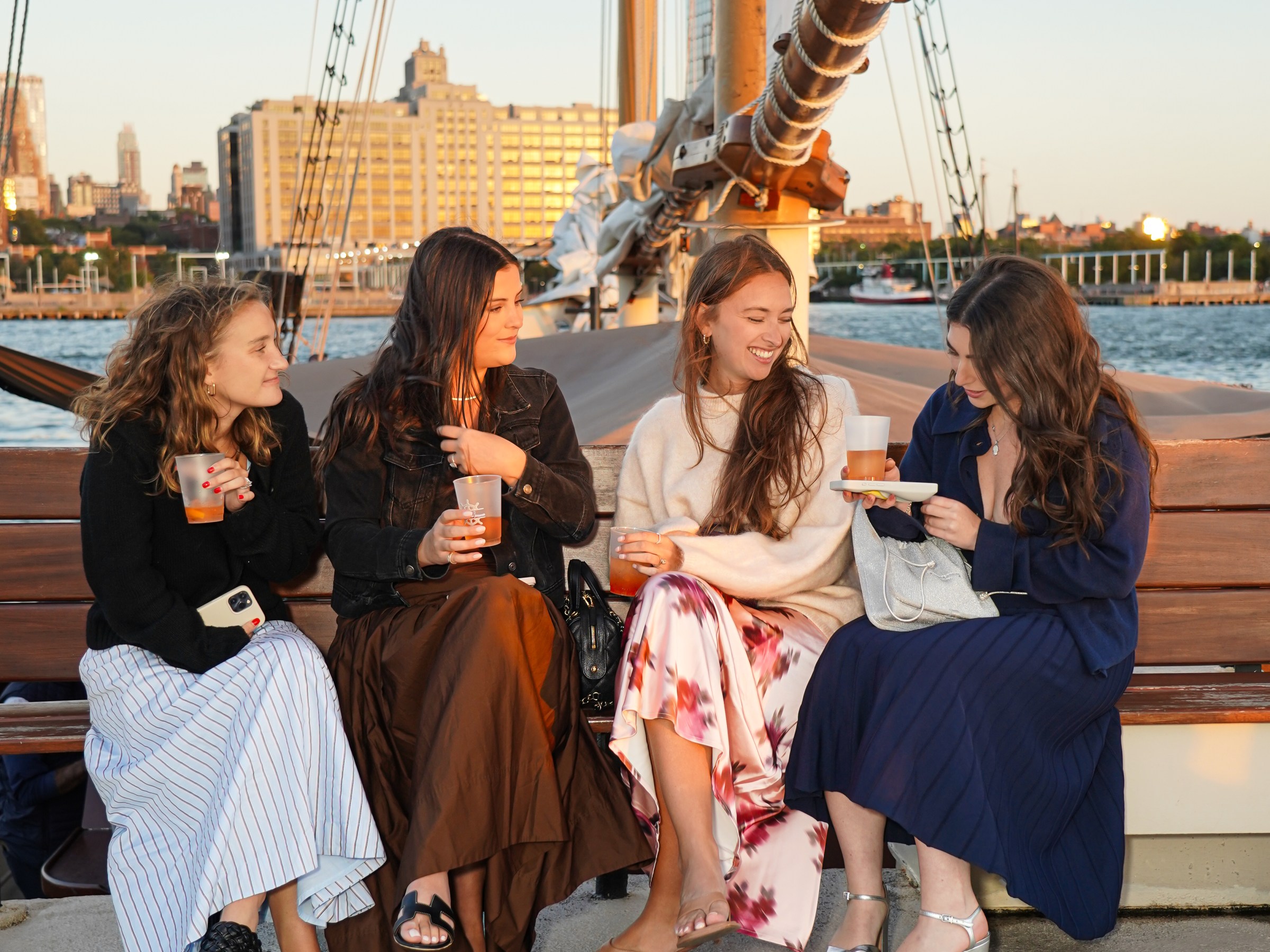 Four women sit on a boat deck at sunset, holding drinks and smiling.