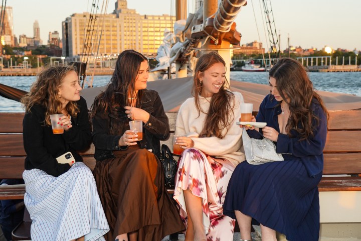 Four women sit on a boat deck at sunset, holding drinks and smiling.