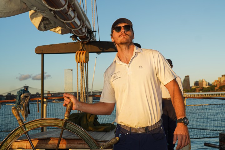 Man in sunglasses steering a sailboat at sunset with city skyline in background.