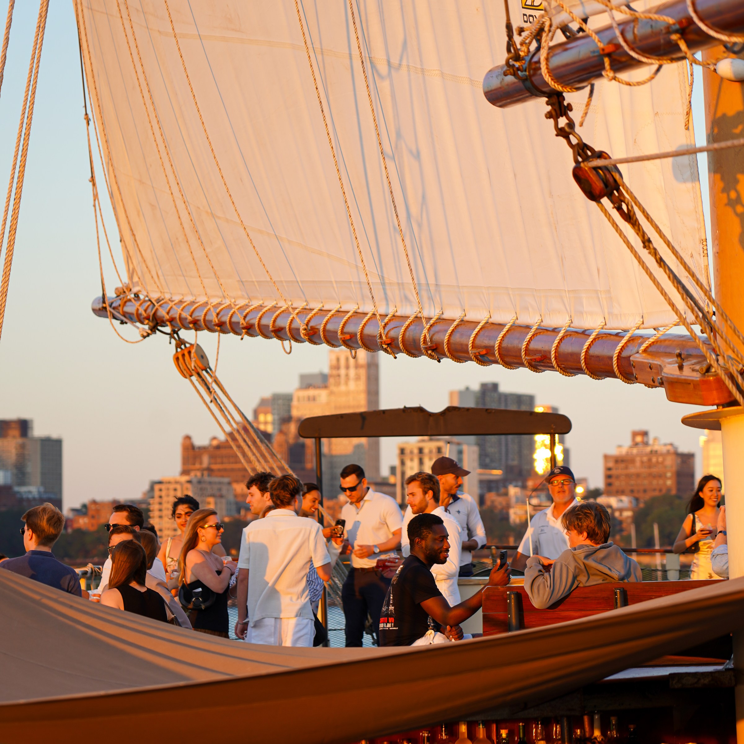 People socializing on a sailboat with large sails at sunset, cityscape in the background.