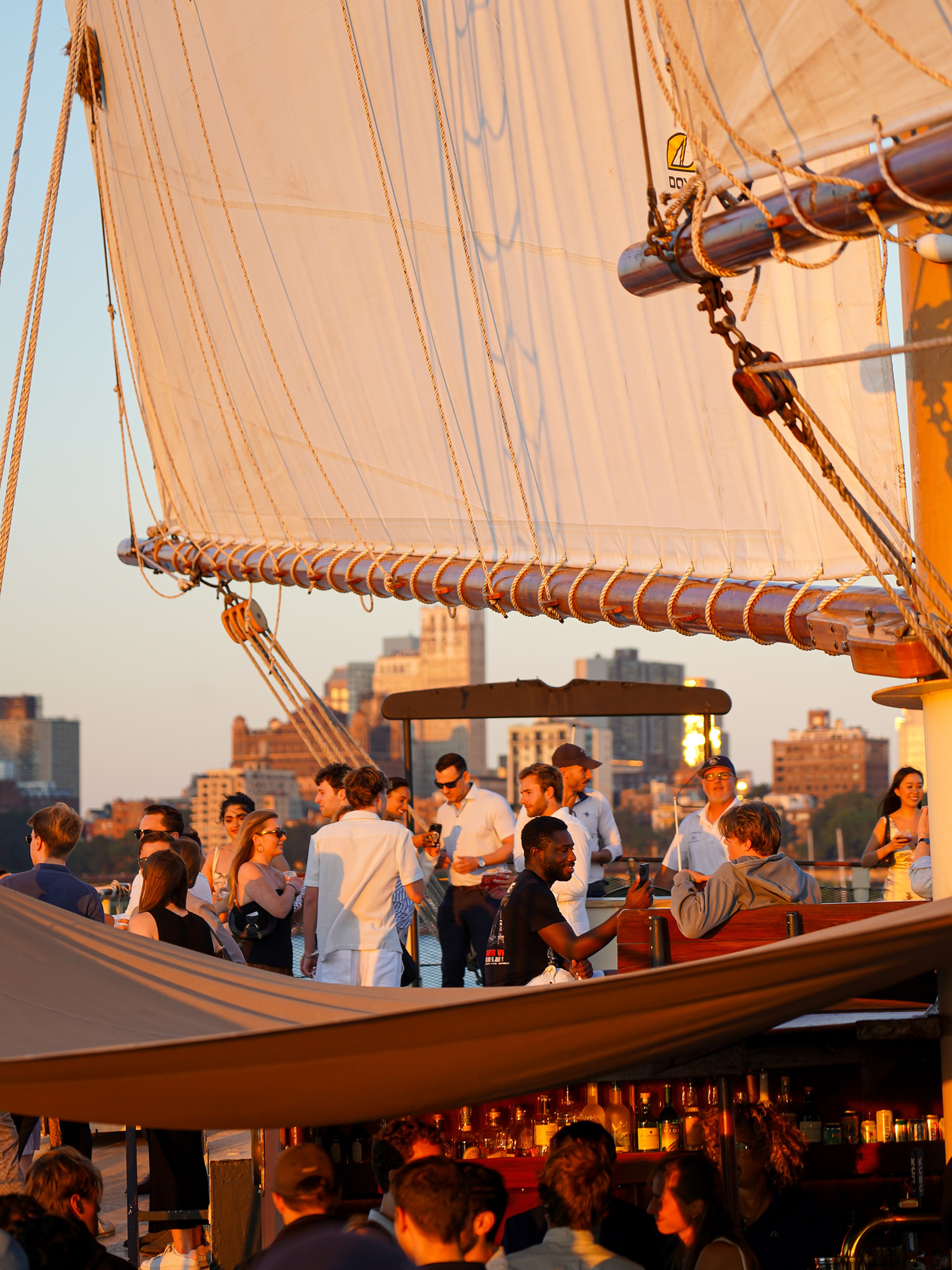 People socializing on a sailboat with large sails at sunset, cityscape in the background.