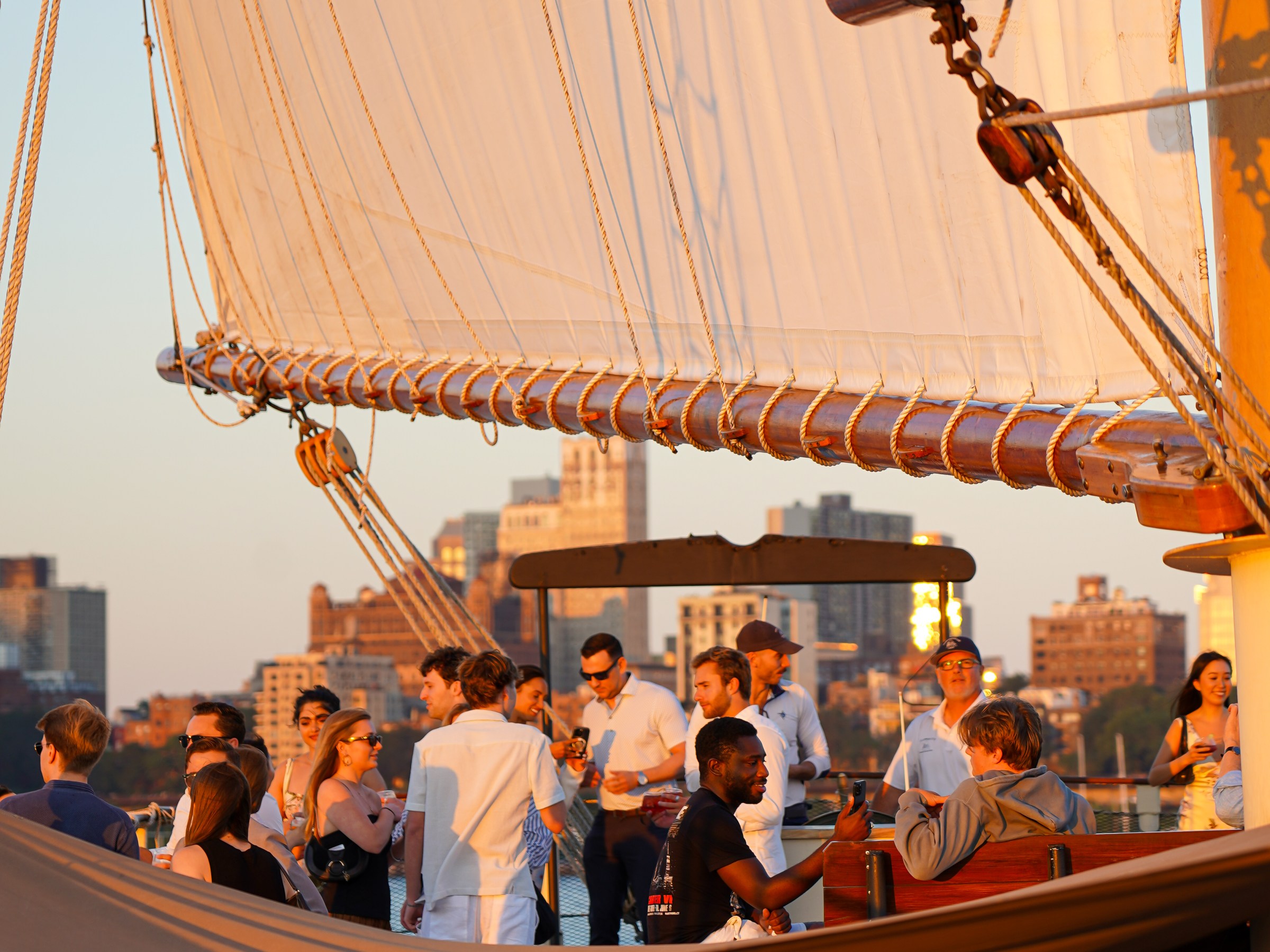 People socializing on a sailboat with large sails at sunset, cityscape in the background.