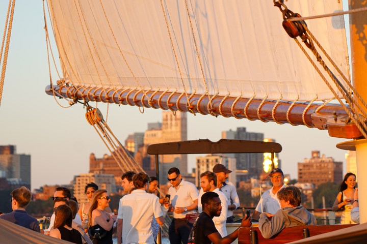 People socializing on a sailboat with large sails at sunset, cityscape in the background.