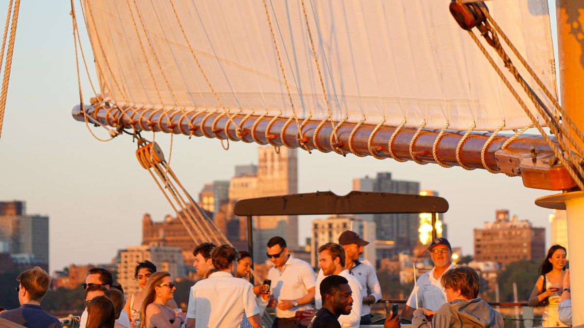 People socializing on a sailboat with large sails at sunset, cityscape in the background.