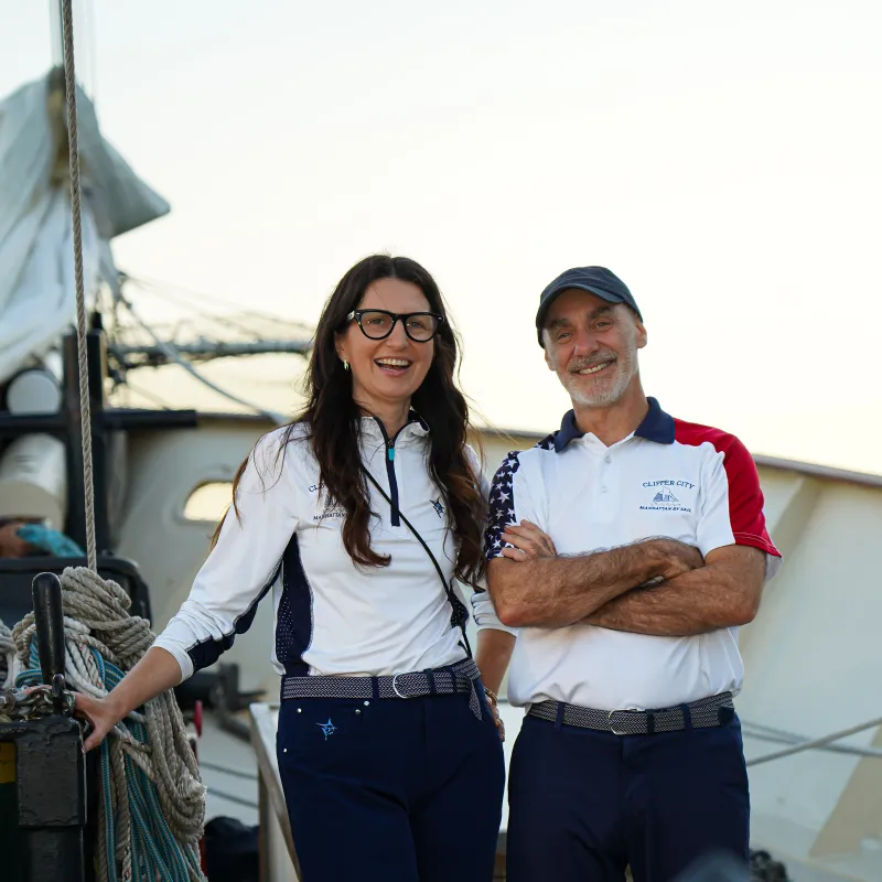 Two people smiling on a boat deck with ropes and sails visible.
