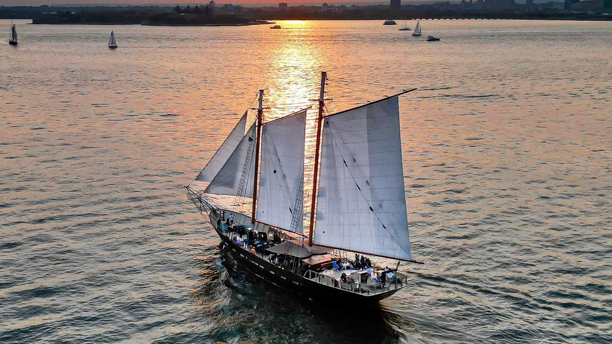 Sailing ship on calm water at sunset with city skyline in background.