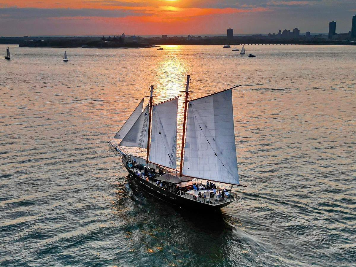 Sailing ship on calm water at sunset with city skyline in background.