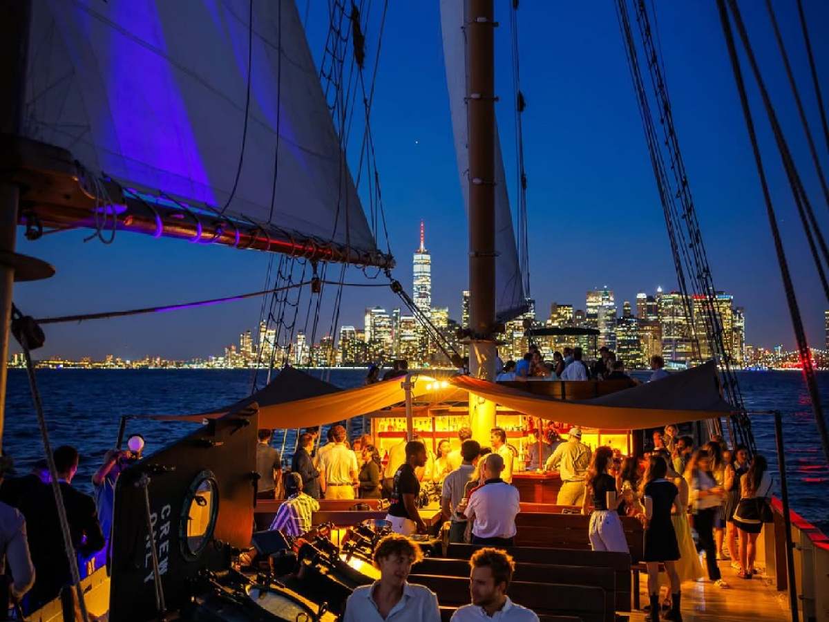 Sailboat with people at night, city skyline in background, lit by warm lights.