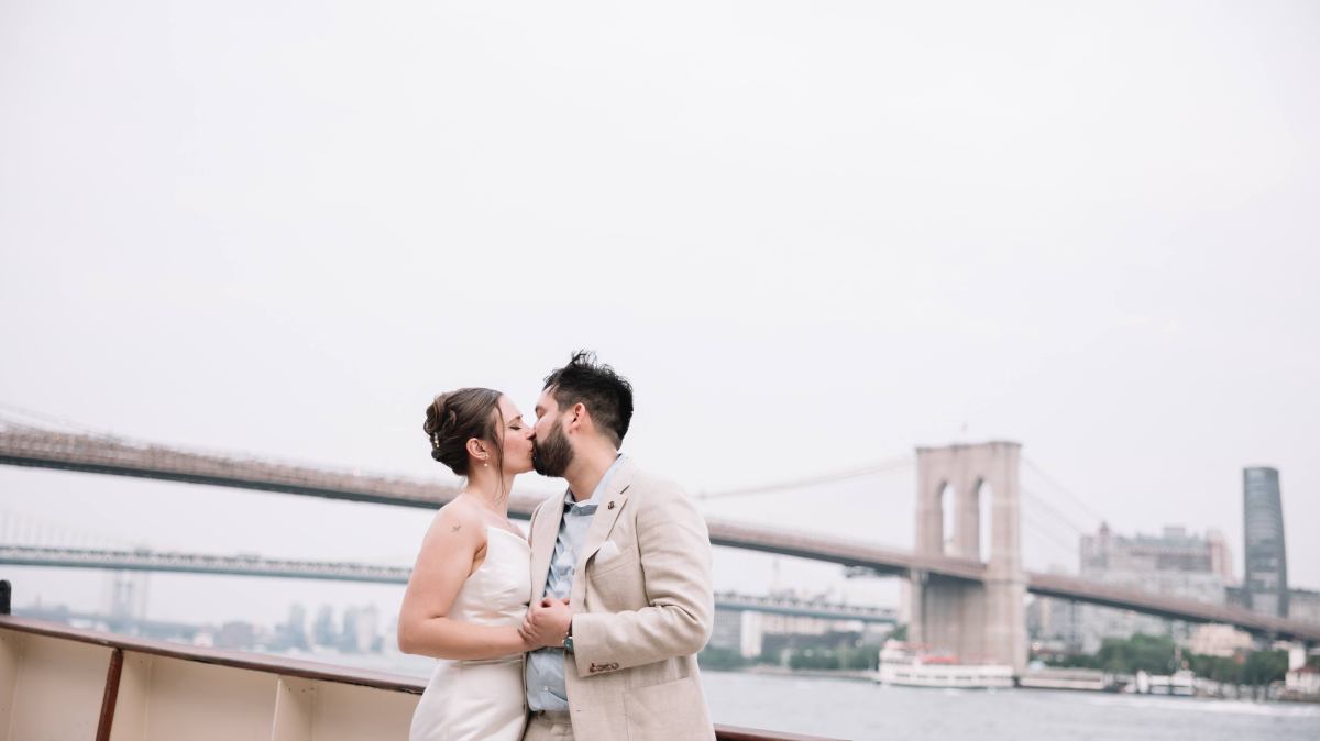 Couple in wedding attire kissing on a boat with a bridge in the background.