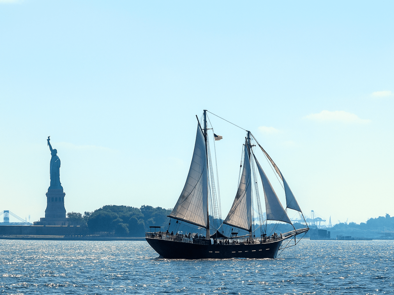 Sailboat on water with Statue of Liberty visible in the background under a clear blue sky.
