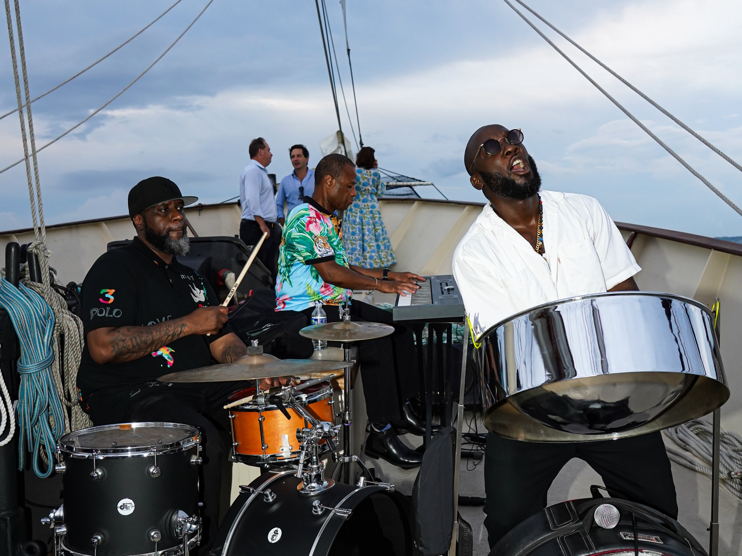 Band playing on a boat with drums, keyboard, and steel pan under cloudy sky.