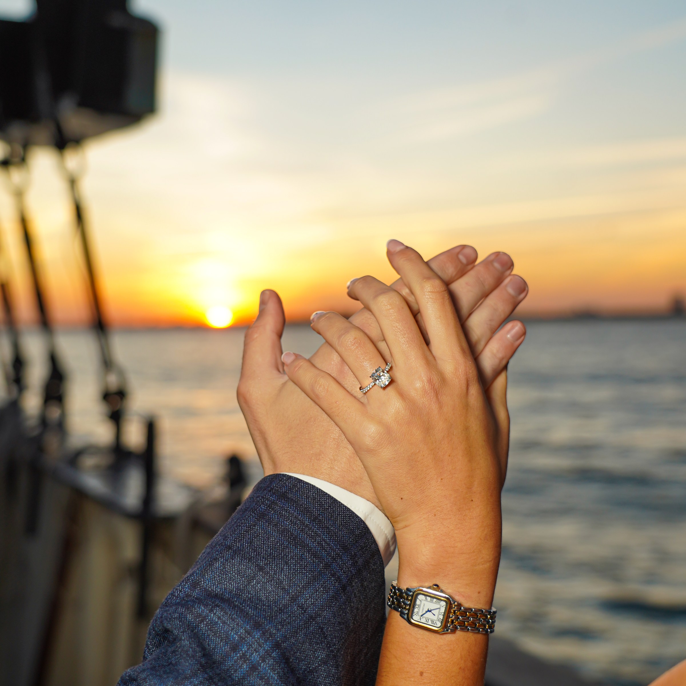 Hands with an engagement ring raised against a sunset over water.