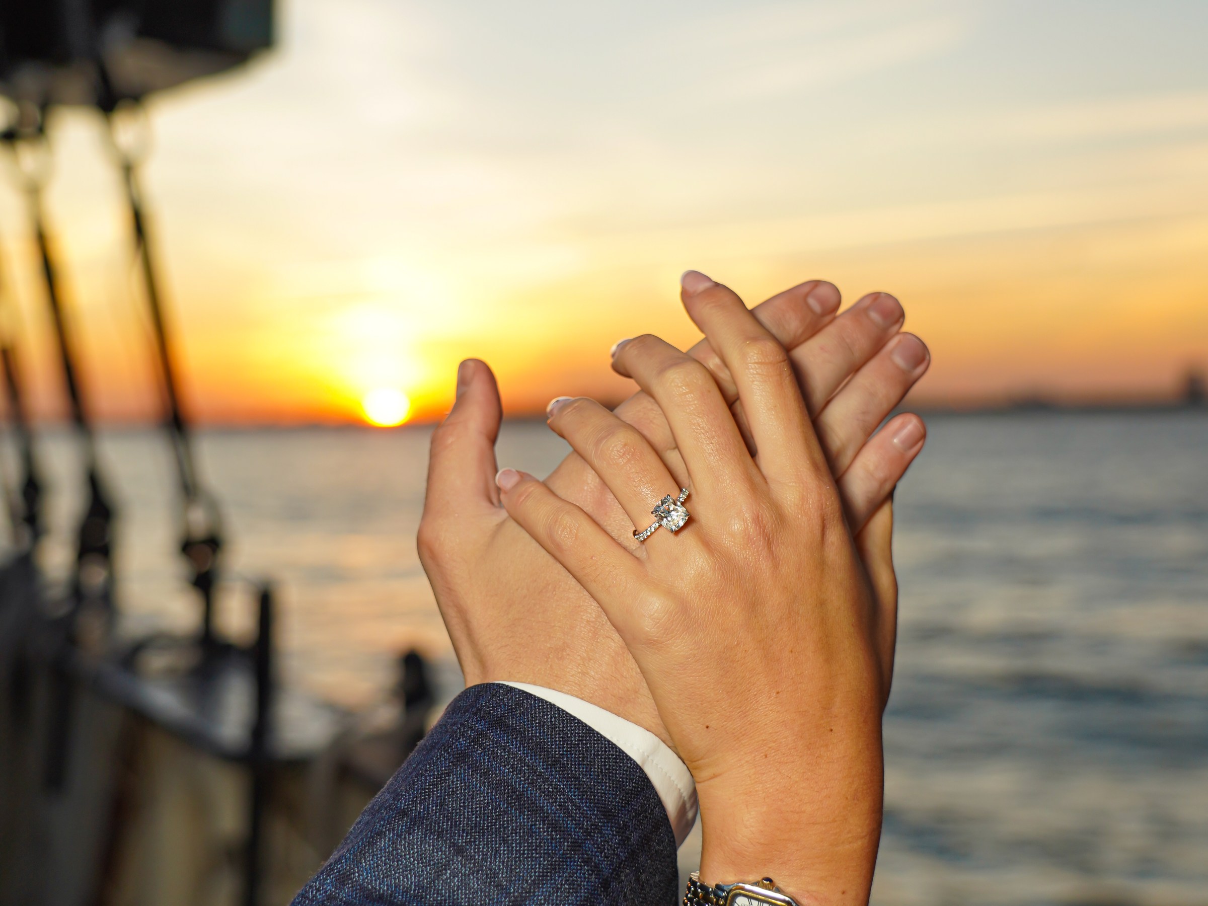 Hands with an engagement ring raised against a sunset over water.