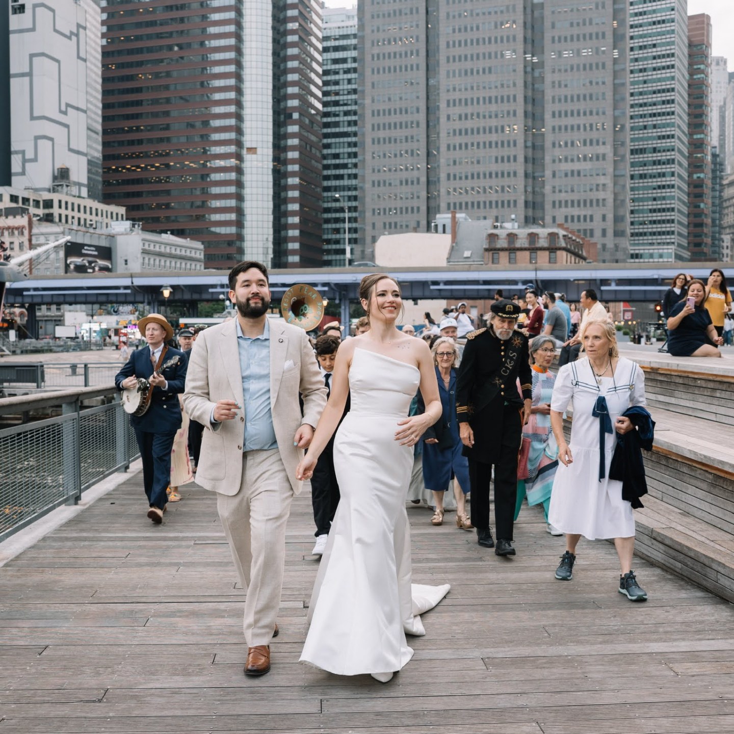Bride and groom walking on a boardwalk with a crowd, cityscape background.