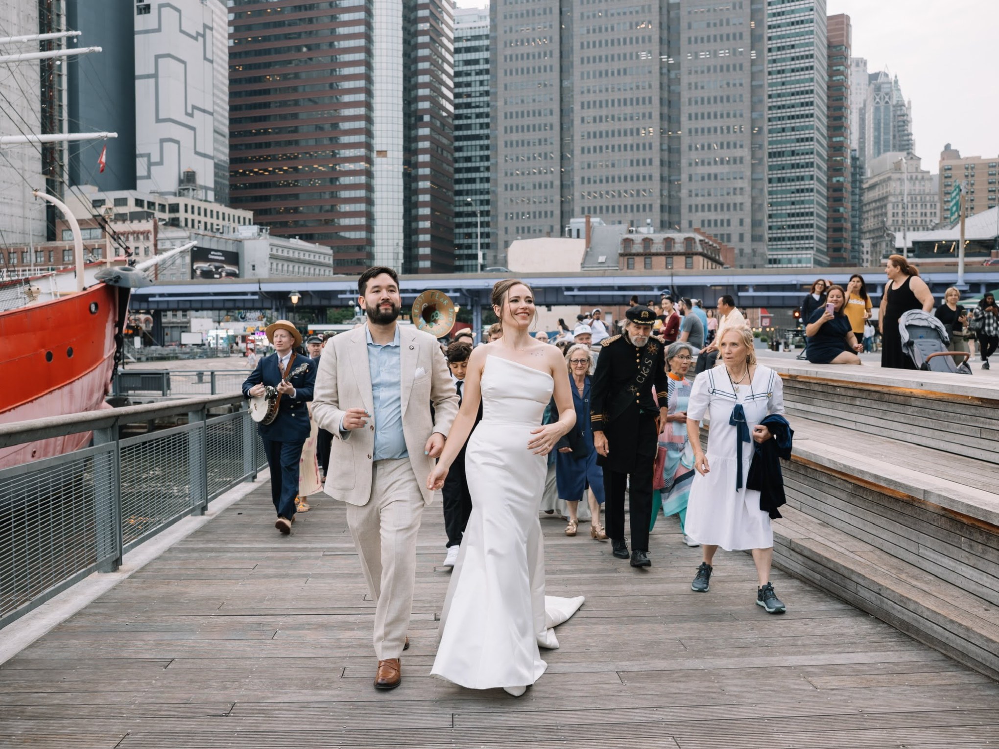 Bride and groom walking on a boardwalk with a crowd, cityscape background.