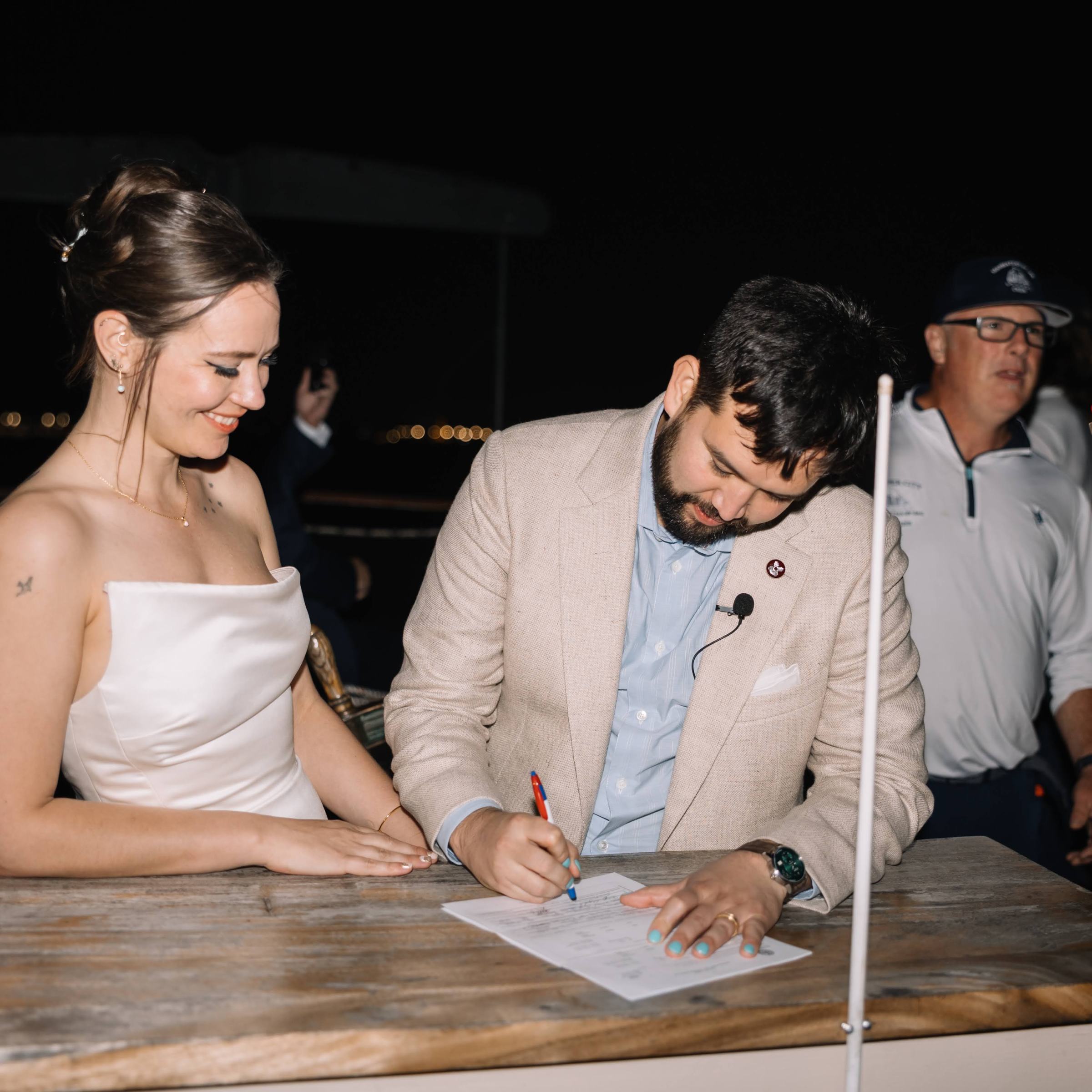 Couple signing a document on a boat at night with onlookers nearby.