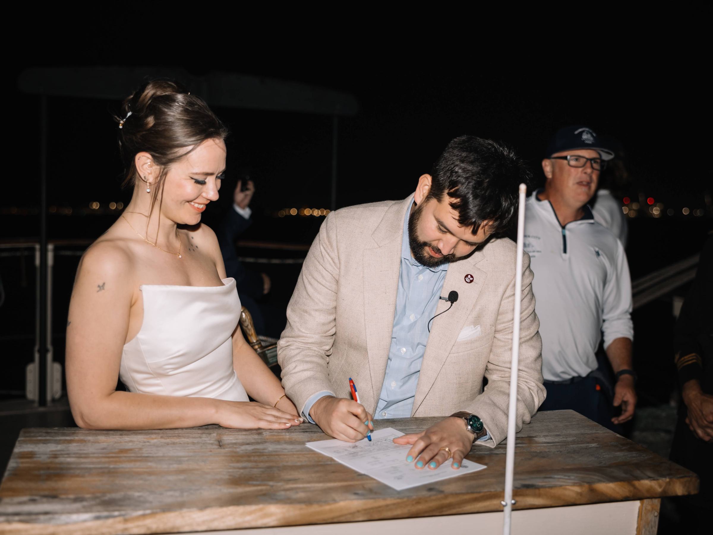 Couple signing a document on a boat at night with onlookers nearby.