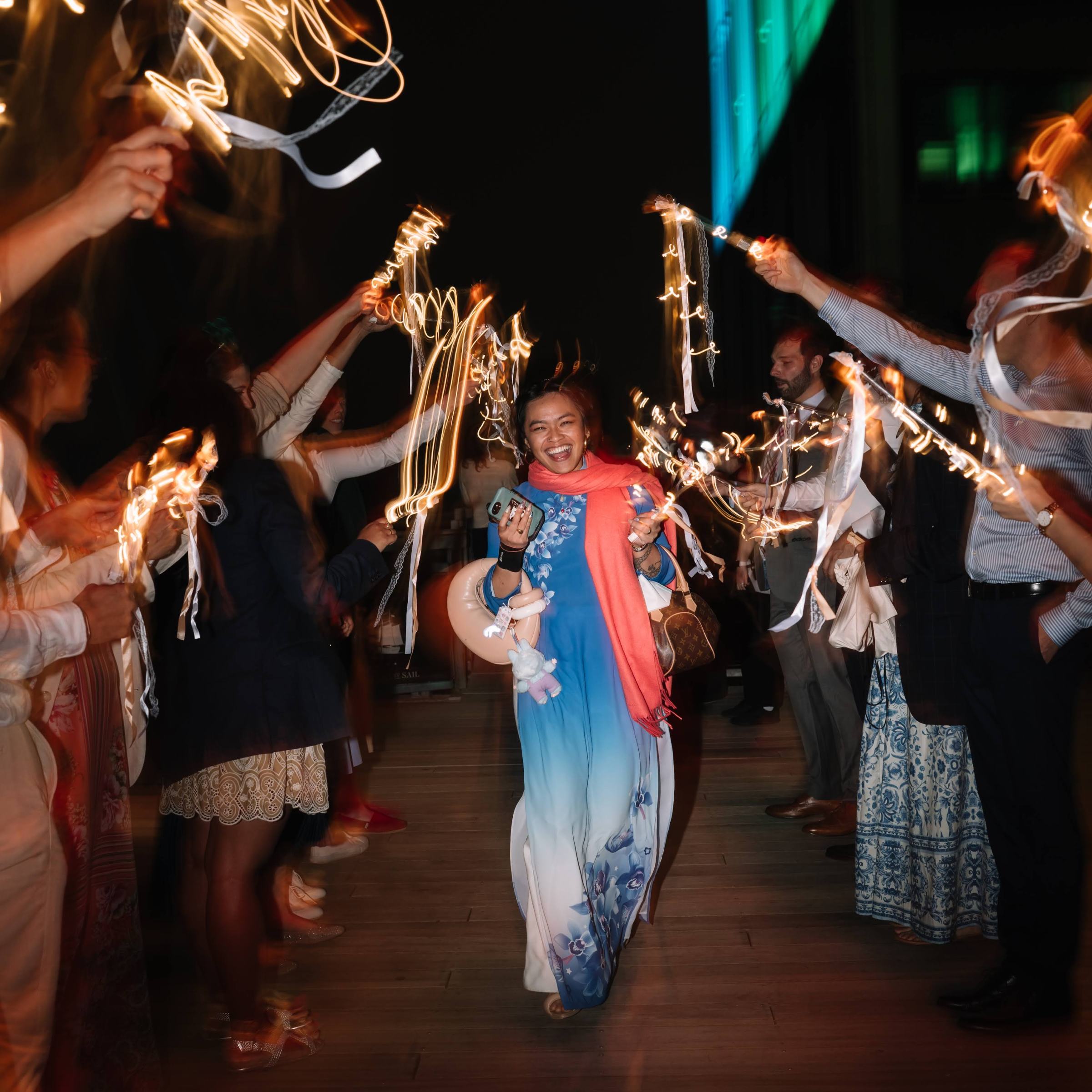 Woman walking through a tunnel of people holding sparklers at night, wearing a colorful dress.