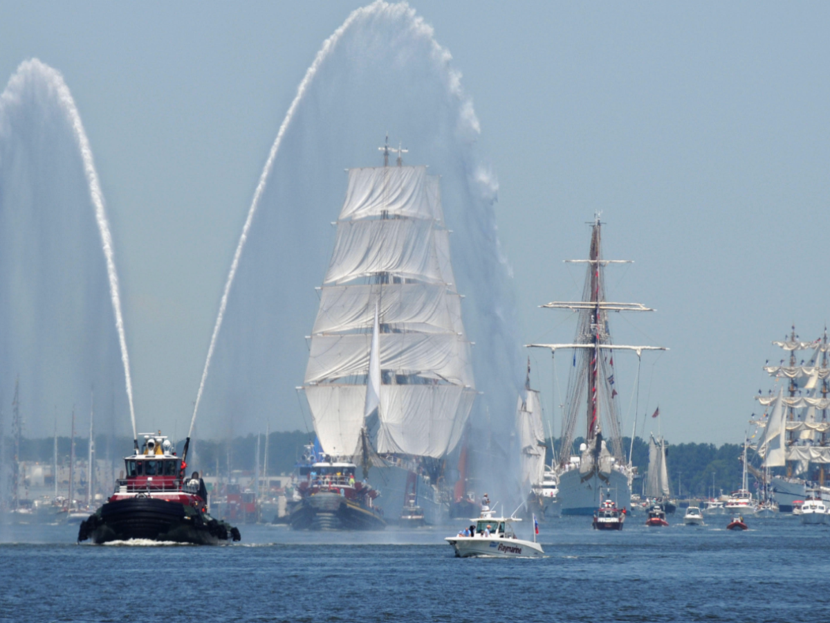 Tugboat spraying water near tall ships with sails on a calm sea.
