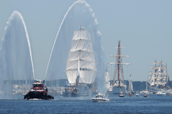 Tugboat spraying water near tall ships with sails on a calm sea.
