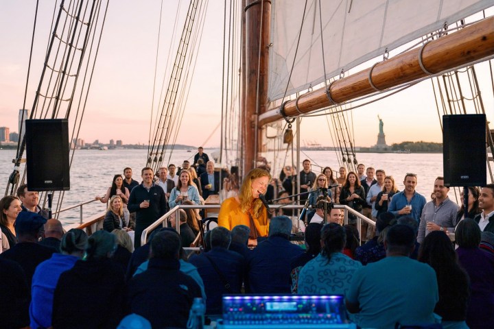 Woman performing on a sailboat with a crowd, sunset, and the Statue of Liberty in the background.