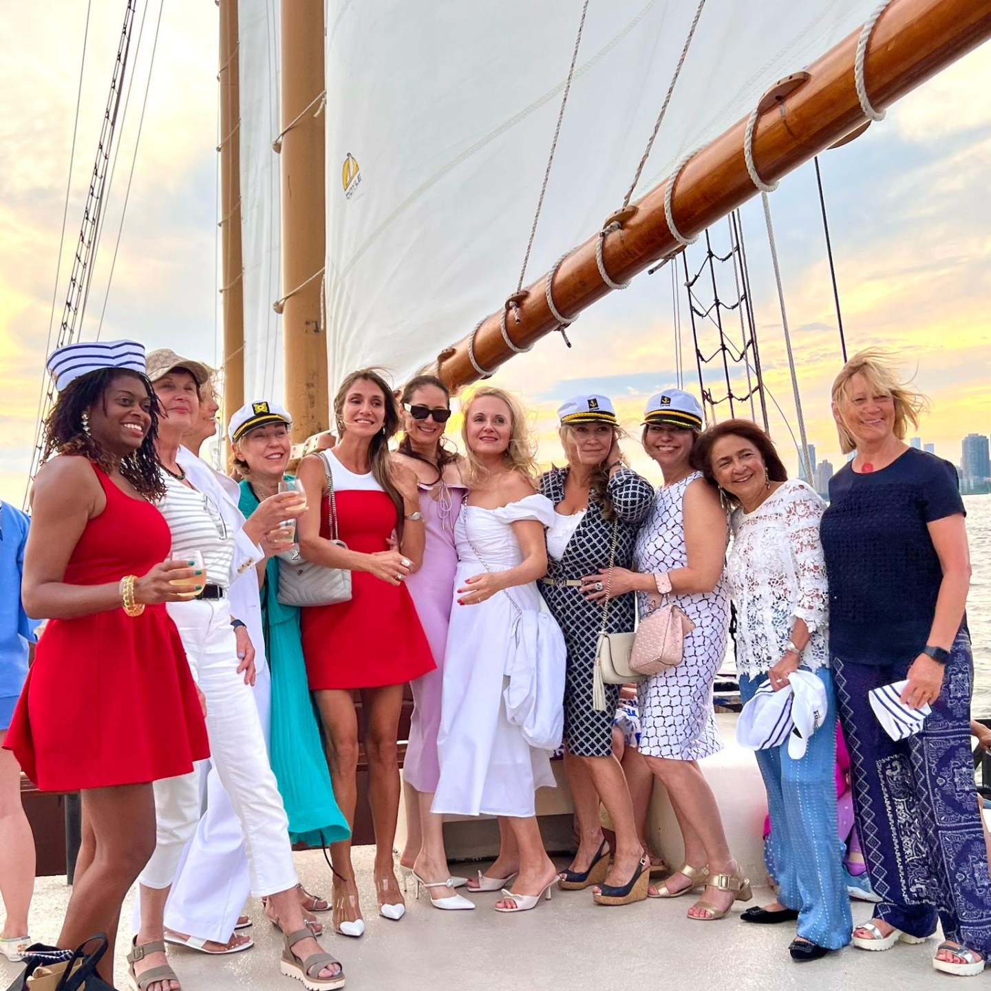 Group of women posing on a sailboat deck at sunset, wearing summer dresses and sailor hats.