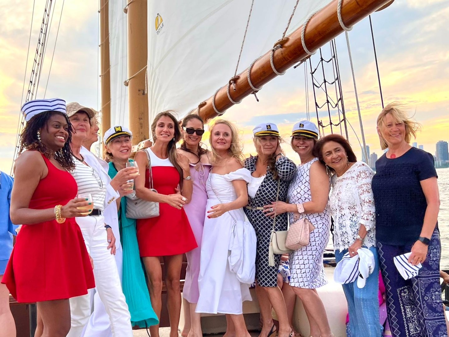 Group of women posing on a sailboat deck at sunset, wearing summer dresses and sailor hats.