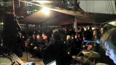 People gathered under a tent on a ship deck at night with a drummer visible.
