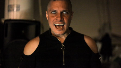 Person with intense expression and glowing eyes in dark setting, wearing a black outfit with shoulder cutouts.