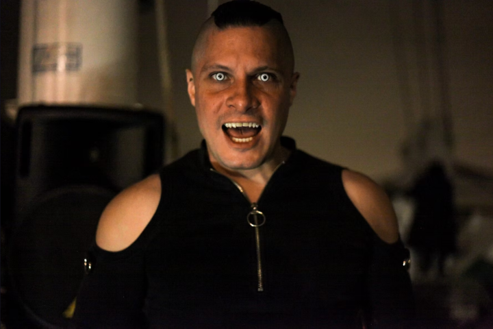 Person with intense expression and glowing eyes in dark setting, wearing a black outfit with shoulder cutouts.