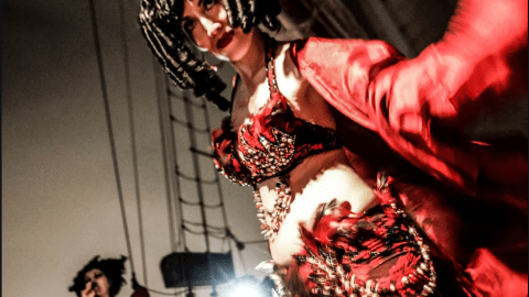 Woman in red costume and wig dances on stage; another in background with microphone.