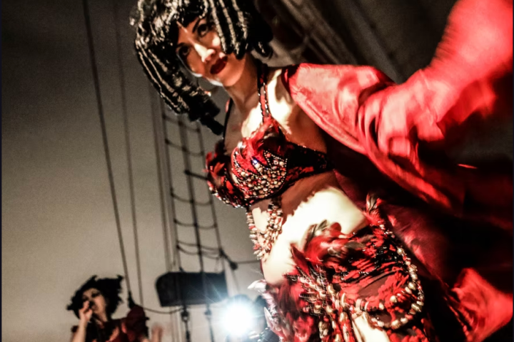Woman in red costume and wig dances on stage; another in background with microphone.