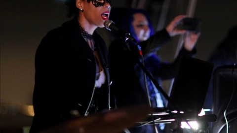 Person in a mask singing and playing keyboard with microphone on stage.