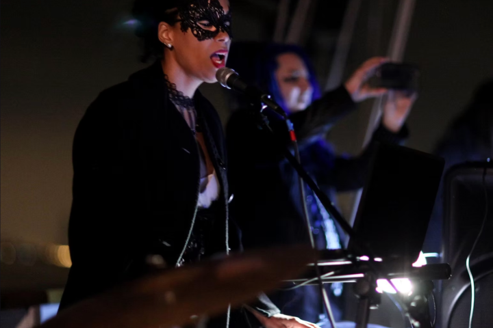 Person in a mask singing and playing keyboard with microphone on stage.