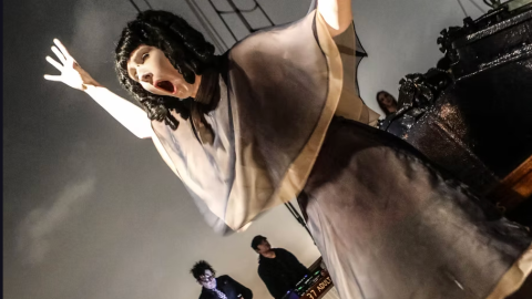 Performer in costume with outstretched arms, theatrical pose on ship deck, musicians in background.