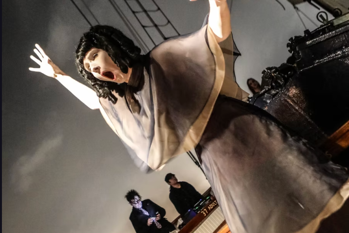 Performer in costume with outstretched arms, theatrical pose on ship deck, musicians in background.