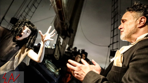 Performer with mime makeup gestures dramatically at a man in a suit on a ship's deck.